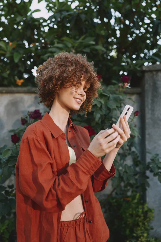 Cheerful woman outdoors, smiling while using a smartphone, wearing a rust jacket, casual lifestyle moment, natural light, relaxed posture, lush greenery and garden backdrop
