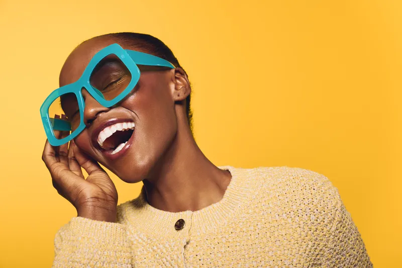 Happy African woman with dark skin wearing large turquoise sunglasses and a yellow sweater smiles with closed eyes on bright yellow background. Concept of joy, fashion, and summer style.