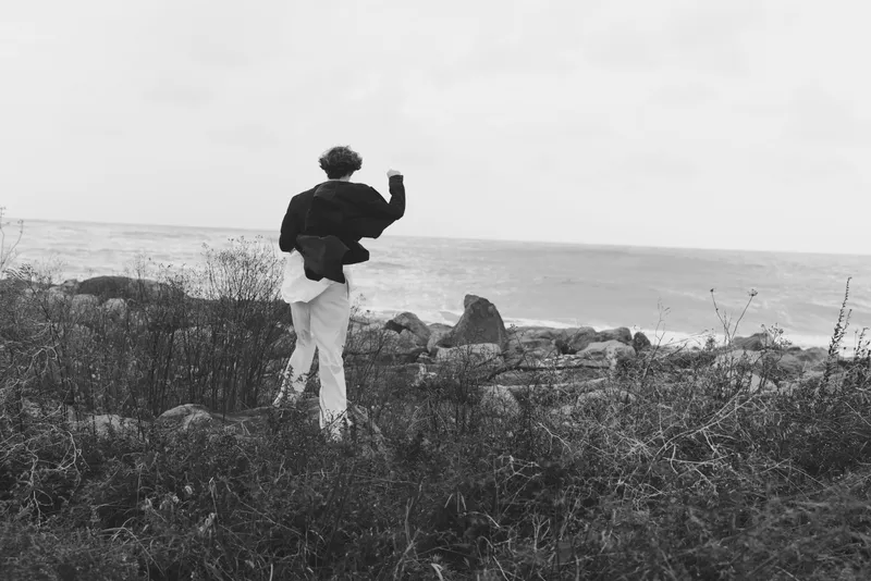 Silhouette of a lone figure walking along a grassy coastline toward the sea on a windy day, a black and white scene highlighting rocks, surf, and vast horizon.
