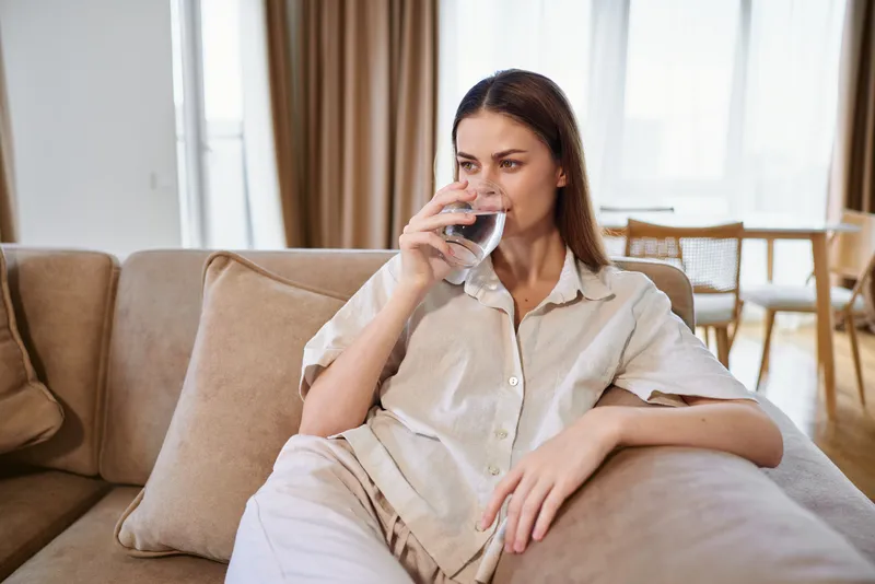 Woman sitting on sofa in livingroom drinking water from glass at home, casual portrait showing relaxation and calm, loungewear comfort and healthy hydration in interior sunlight.