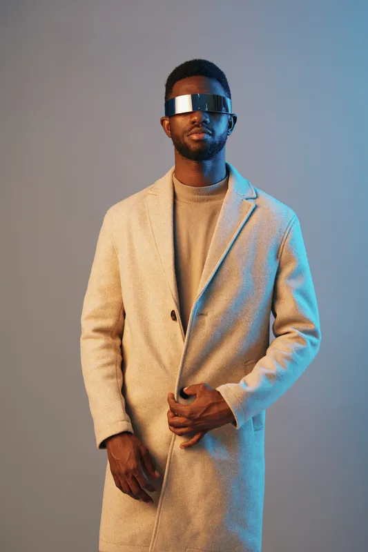 Stylish black male model wearing a modern, light colored coat and futuristic sunglasses, exuding confidence against a soft blue backdrop