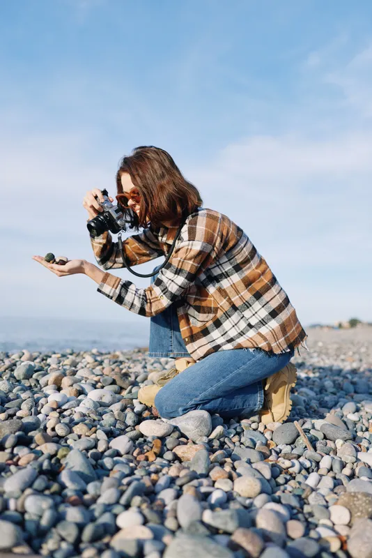 Photography of a photographer kneeling on a pebble beach with a camera, capturing candid moments amid outdoor landscape, nature and focused composition.