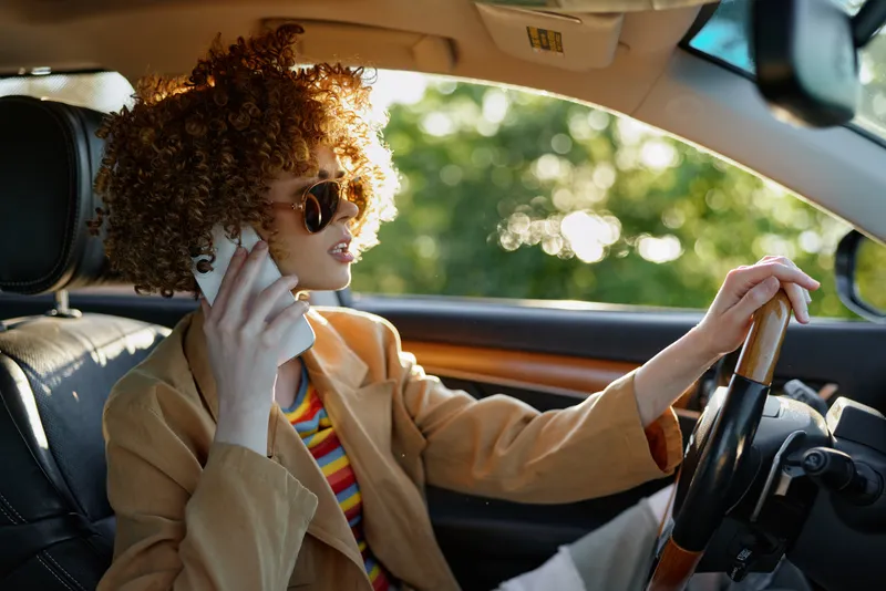 Young woman with curly hair drives a car in warm retro film color, wearing a beige coat and striped top, smiling as sunlight streams through the window, capturing freedom and nostalgia.