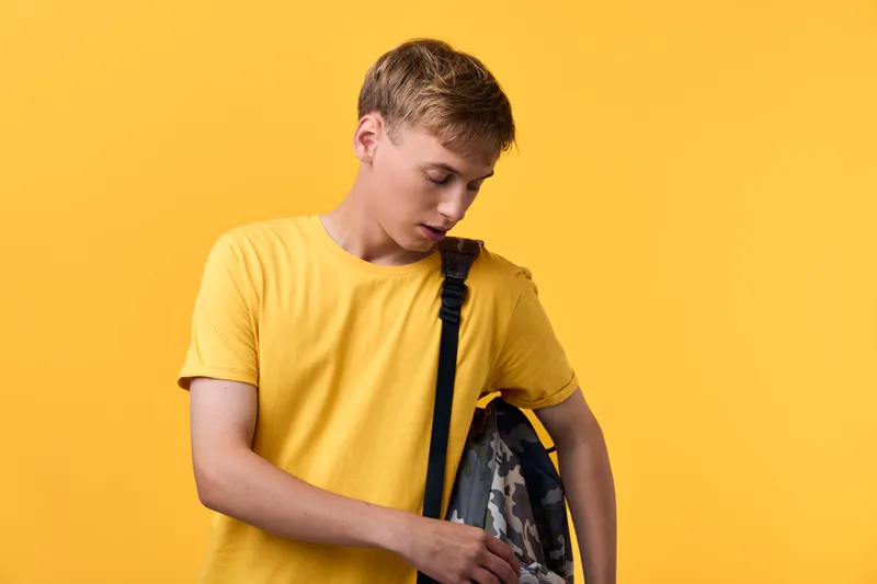 Student wearing a yellow shirt with a backpack stands against a bright yellow background, suggesting casual school attire, everyday travel and youthful preparation for classes.