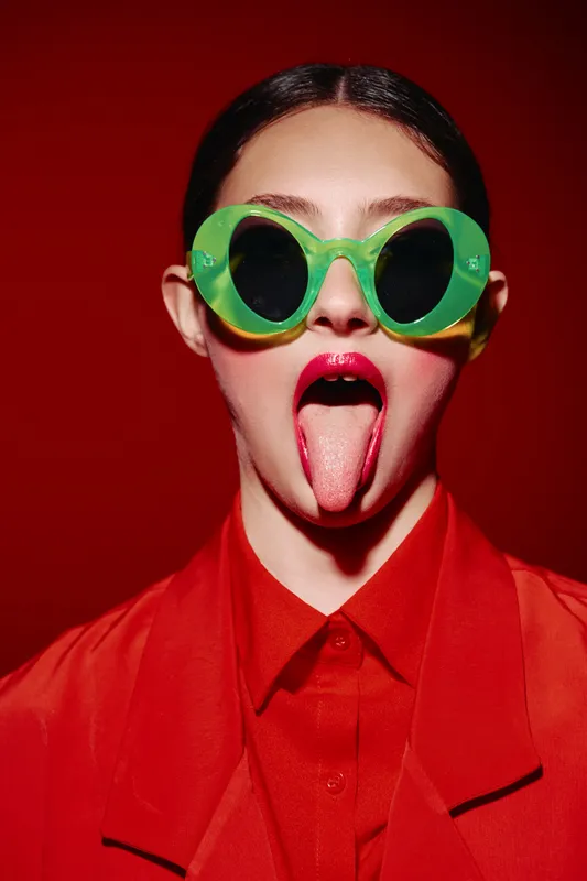 Fashion model with glossy lipstick and striking green sunglasses poses on a red background sticking out tongue. Creative beauty look with contrasting colors captures daring expression.