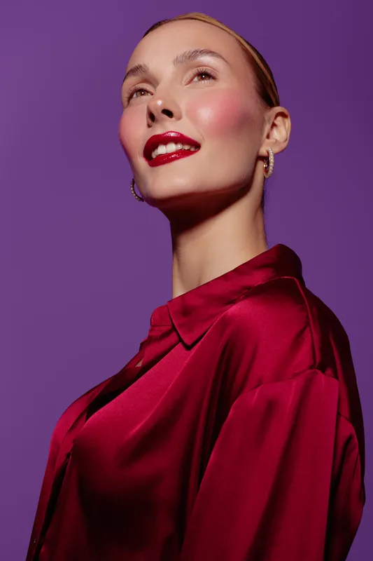 Confident woman with red lipstick and shiny satin blouse poses against purple background. Glamorous makeup and elegant earrings highlight her sophisticated style and radiant expression.