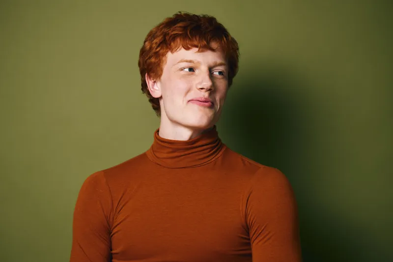 Young man with curly red hair wearing brown turtleneck looking to side with slight smile against green background. Studio portrait with casual confident expression and simple clothing.