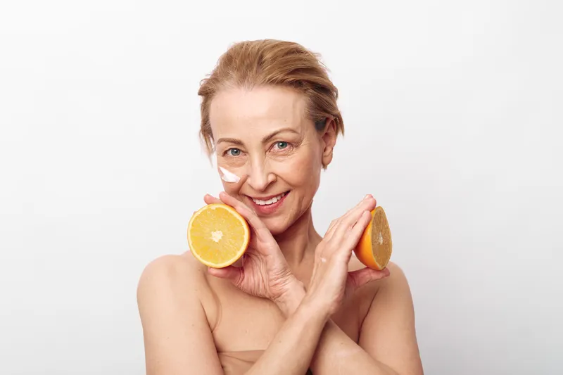 Smiling mature woman with natural skin holding two orange halves near her face. Concept of healthy lifestyle, vitamin c, skincare, and natural beauty care on white background.