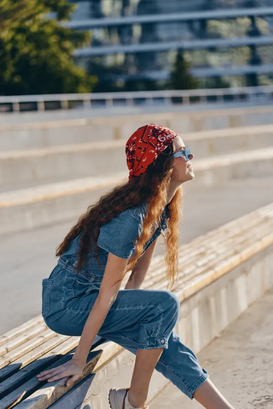 Lifestyle woman in modern boho-western denim street style sits outdoors wearing a red bandana and sunglasses, showing relaxed and stylish summer vibes in film color tones.