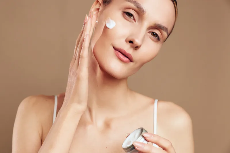 Young woman applies moisturizer cream on her face with hand holding jar isolated on beige background natural skincare beauty routine closeup portrait