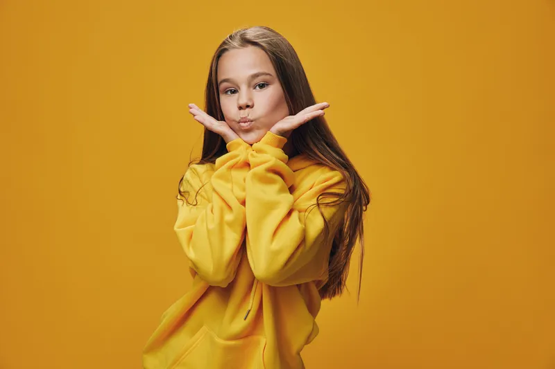 Girl in yellow hoodie posing with hands near face on yellow background, looking playful and confident with long hair and casual clothing in studio portrait lighting.