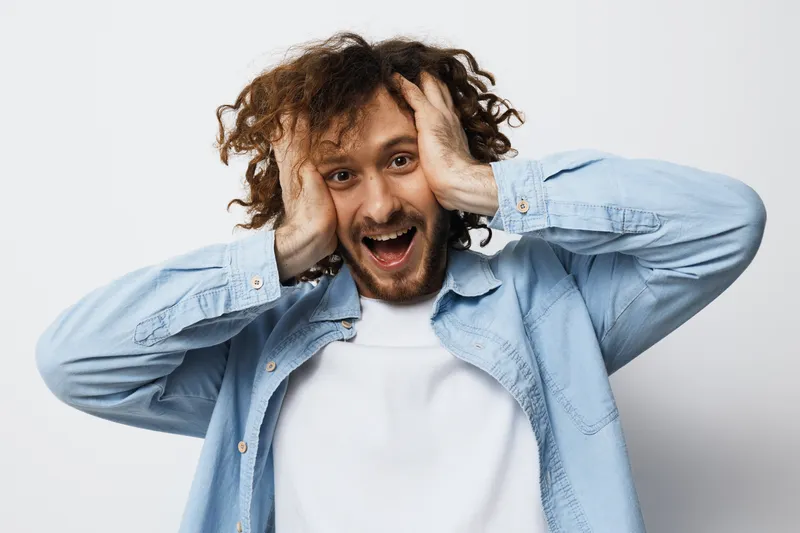 Young joyful man with curly hair expressing surprise and excitement against a minimalistic background wearing a light blue shirt, perfect for emotions related to happiness