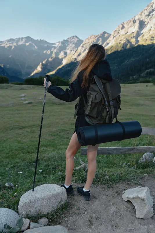 A lone hiker stands on a rocky trail in a mountain meadow, gripping a trekking pole. She wears a sturdy backpack with a rolled mat, preparing for outdoor exploration and enjoying wide panoramic