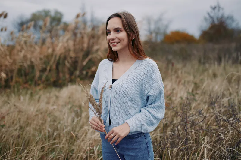 Young woman in a natural field wearing a cozy sweater, smiling and holding dried grasses, captured with authentic and credible lifestyle portraiture and warm candid expression.