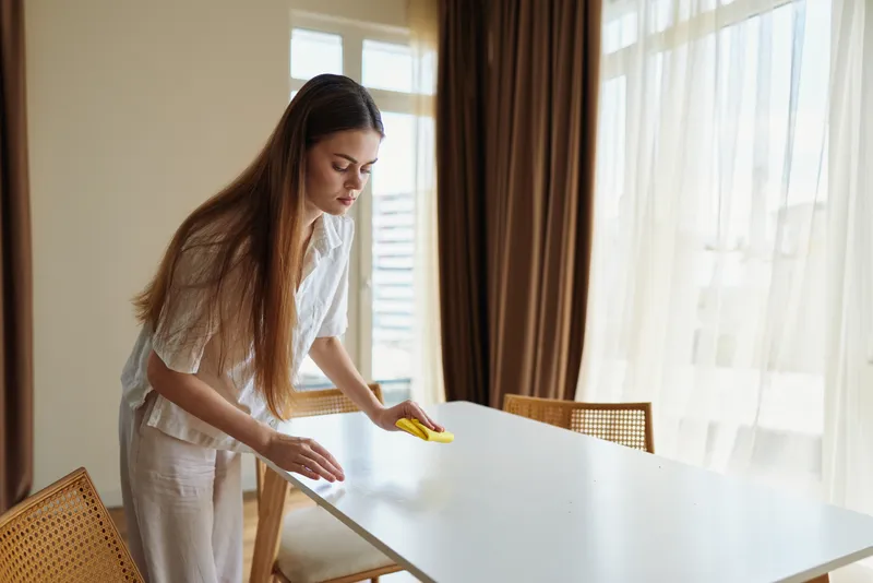 Woman cleaning dining table with sponge and cloth in bright home interior by window, wiping surface for hygiene and tidiness in casual domestic scene with simple furniture.