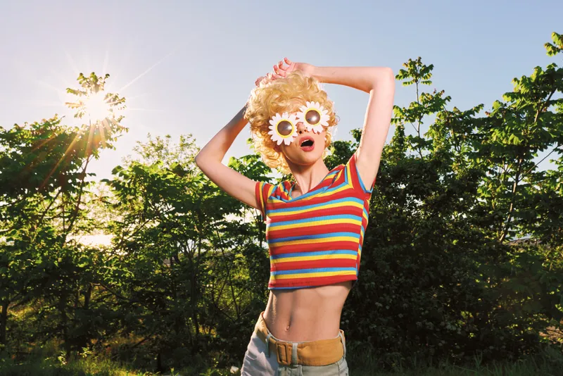 Energetic young woman dances outdoors in a striped crop top and flower sunglasses, sunlit joy and a breezy park vibe