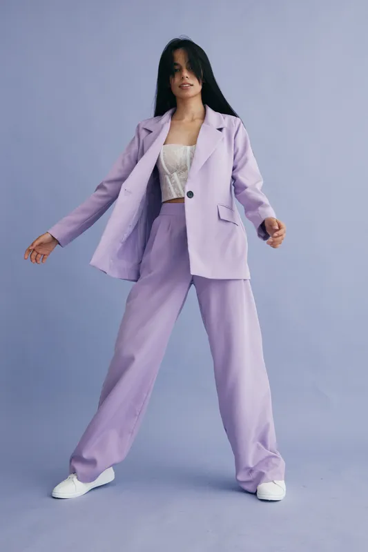 young woman in lilac pantsuit posing confidently against a pastel blue background, wearing white sneakers and a white lace top, studio fashion portrait style