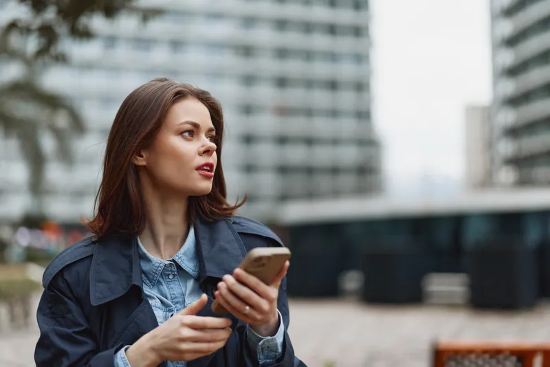 Young woman with brown hair holds a smartphone and looks away thoughtfully while sitting outdoors in an urban environment wearing a dark jacket and denim shirt.