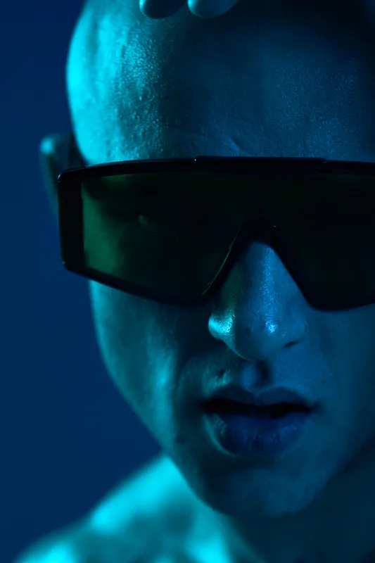 Close-up of a serious man wearing futuristic sunglasses, highlighting strong facial features and intense expression under blue light. Concept: modern masculinity.