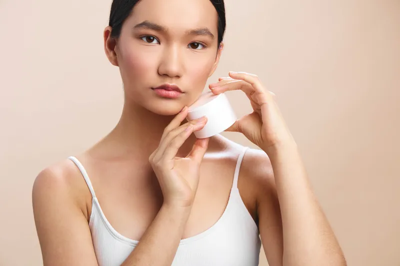 Asian woman applies cream from a small jar in a clean studio setting. She looks softly at the camera, conveying calm wellness and intimate self care in a minimalist environment.