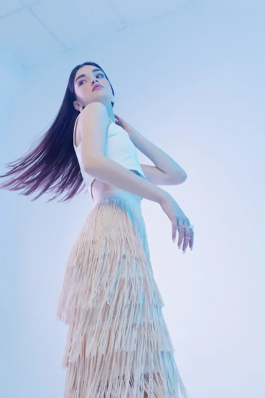 Young woman in a white top and fringed skirt poses with flowing hair against a light blue background. Concept of beauty, fashion, elegance, and modern style.