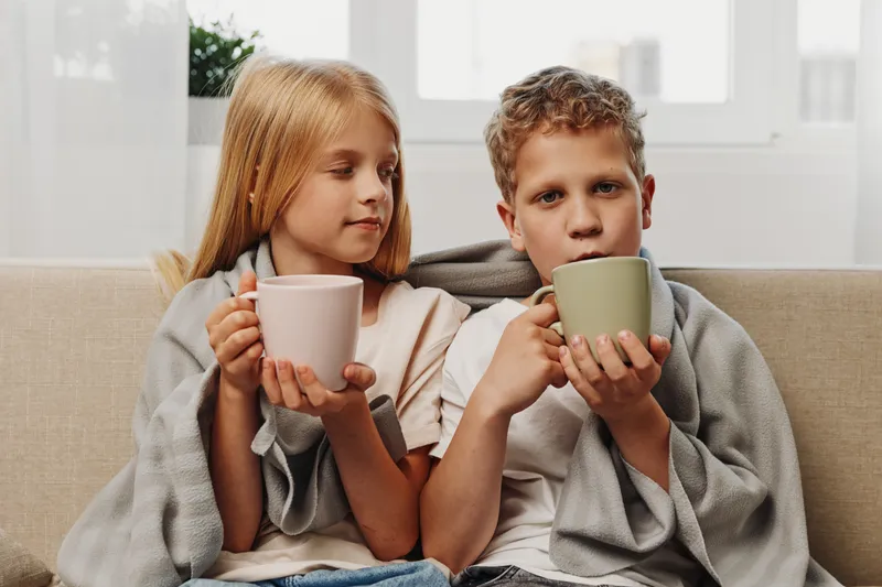 Two children wrapped in a blanket drink hot beverages on a sofa in a bright room. Cozy moment of warmth and comfort at home with warm drinks.