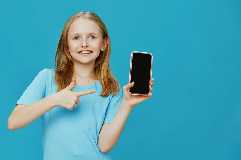 Cheerful young girl pointing at a smartphone against a vibrant blue background, showcasing excitement and friendliness for promotional content