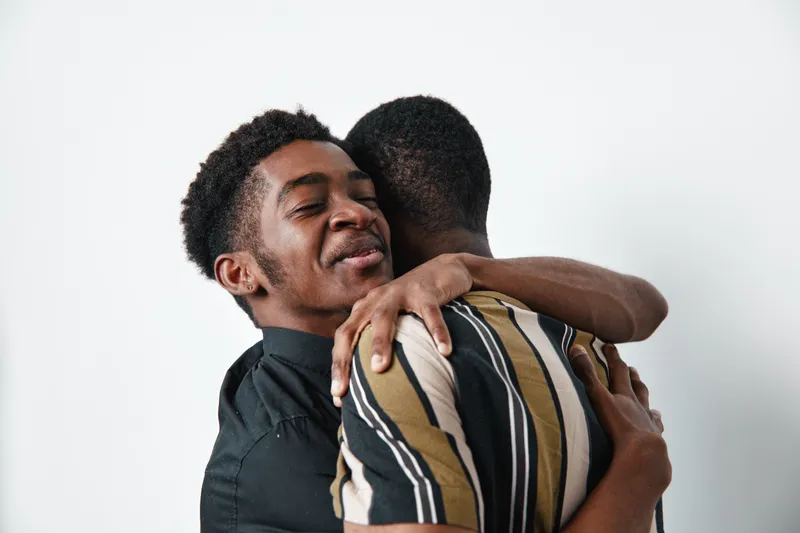 Two young black men hugging each other with eyes closed and smiles, showing friendship and affection in casual clothing on white background. Emotional connection and support.