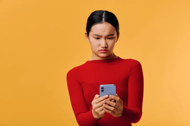Woman in red sweater looking at smartphone with confused expression on orange background. Female holding phone, frustrated face, technology and communication concept with emotions and mobile device.