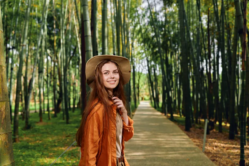 Woman smile hat bamboo path forest longhair portrait: young woman with longhair and straw hat smiles on a sunlit bamboo forest path, wearing autumn jacket and casual outfit for a peaceful nature