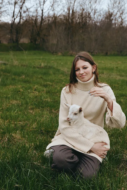 A woman in a cozy sweater sits in a grassy field, gently holding a young goat kid on her lap. A calm lifestyle moment capturing bond with animals and open outdoors.