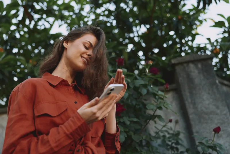 A smiling woman in a rust shirt checks her phone outdoors, with lush greenery in the background, creating a candid lifestyle moment focused on connection and everyday joy