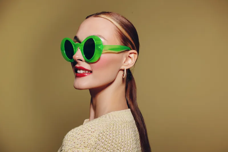 Stylish woman with long hair wears large green sunglasses and red lipstick, posing against beige background. Concept of fashion, beauty, accessories, and confident attitude.
