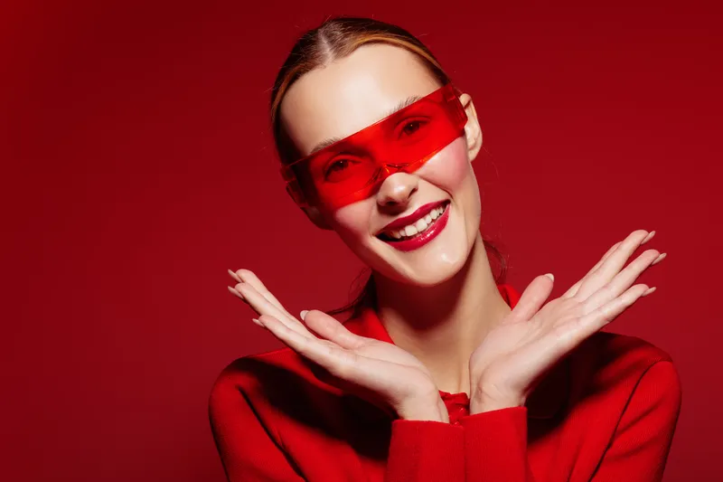 Smiling woman in red sweater and red sunglasses poses with hands near face on red background. Bright studio lighting highlights cheerful mood and vibrant fashion concept.