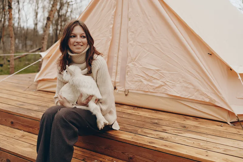 A smiling woman sits on a wooden deck beside a beige camping tent, holding a fluffy white dog. Cozy sweater, soft light, outdoor lifestyle moment that emphasizes companionship and simple camping.