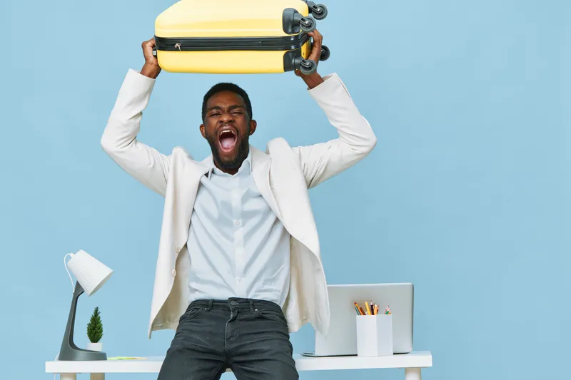 Full body cheerful young black man in white blazer and shirt holding yellow suitcase overhead looking excited joyfully standing indoors against a pastel blue background. People lifestyle concept