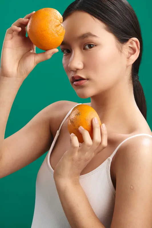 Asian woman holds an orange near her face in a clean studio with a vivid green background, smiling softly. The fresh, modern scene conveys health, vitality, and a light lifestyle mood for stock use.
