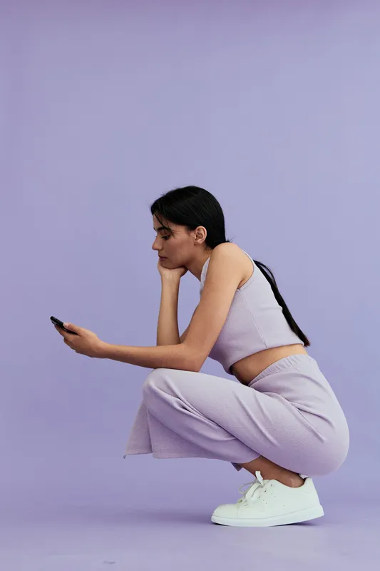 Young woman crouching while looking at smartphone, wearing casual purple outfit and white sneakers against lavender background, focused and relaxed mood.