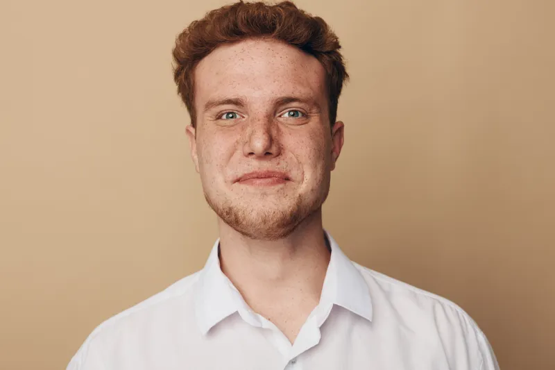 Portrait of a smiling young man with red hair and freckles wearing a white shirt on beige background. Concept of happiness, confidence, and casual lifestyle.