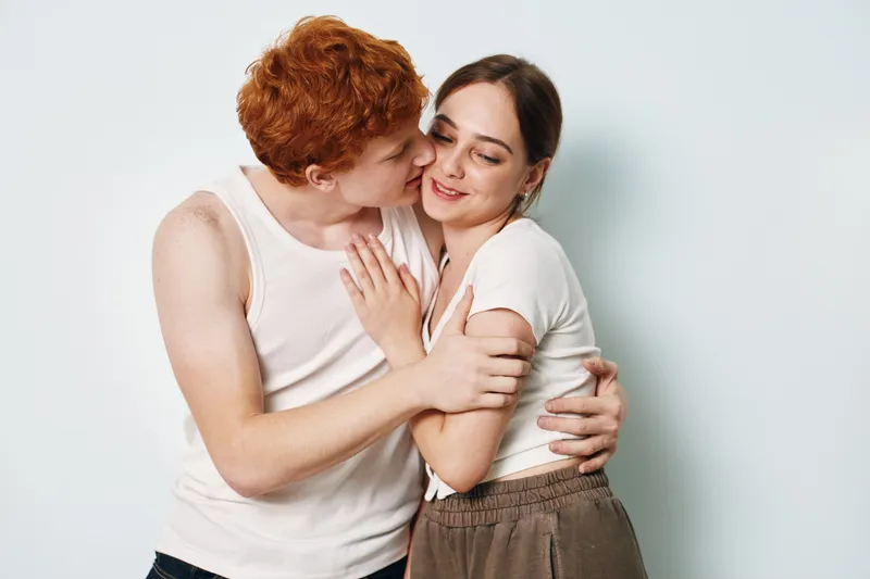 Couple, love, affection, and relationship with young man kissing woman on cheek for romance and intimacy in casual clothes with gentle touch on white background.