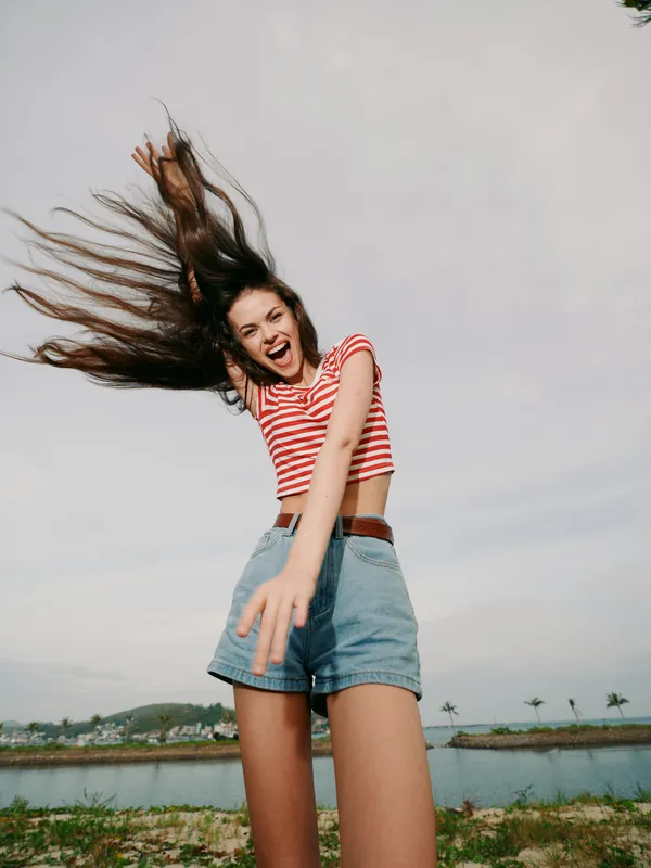 A lively young woman jumps with hair flying and a bright smile, wearing a striped crop top and denim shorts, captured in a carefree outdoor moment by a windy shoreline.
