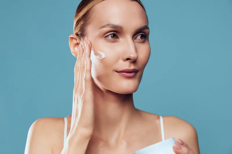 Woman applying white face cream to cheek in a close up portrait, turquoise background, soft studio lighting, concept of skincare, wellness, self care, personal hygiene, calm mood