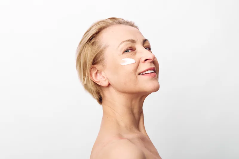 Mature woman with short blonde hair applying face cream on cheek, smiling gently. Beauty concept focused on skincare, anti aging, health, and natural skin care routine on white background.