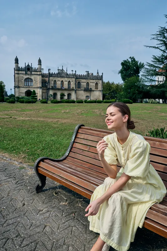 A woman in a light dress sits on a wooden bench in a sunny park, smiling softly while enjoying the scenery, with a historic castle in the background and a peaceful atmosphere