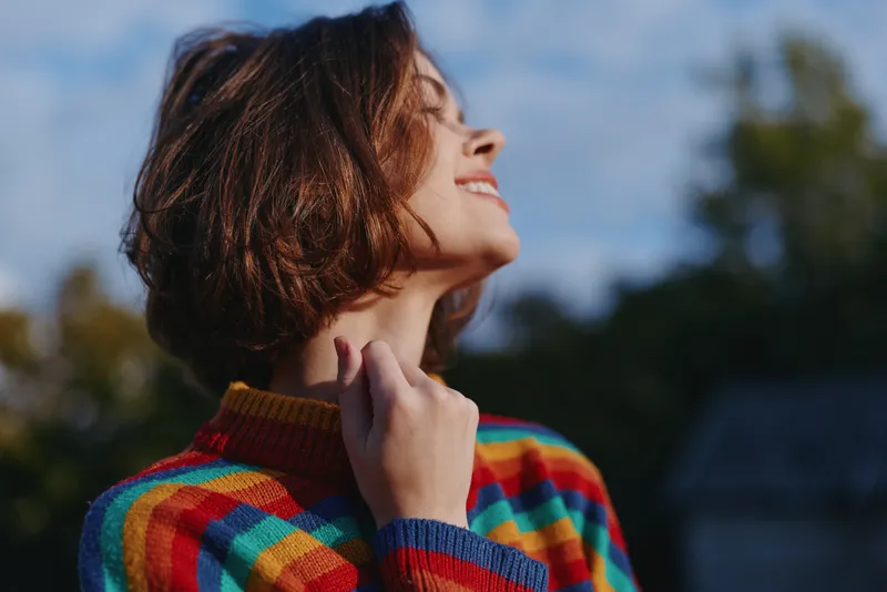 Woman portrait smiling in a colorful sweater outdoors with short hair, profile and sunlight on face, joy visible in expression. Young adult enjoying fresh air and warm natural light.