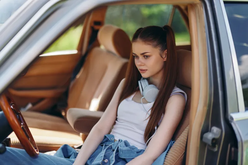 Young woman sitting in a car, wearing headphones, casual outfit, relaxed expression, natural outdoor background, vibrant colors, summer vibe.