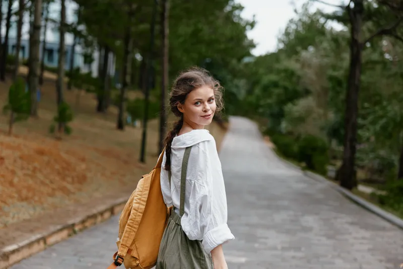 Young woman in a casual outfit walking on a path in a serene park, conveying a sense of freedom and exploration with a joyful expression and a backpack