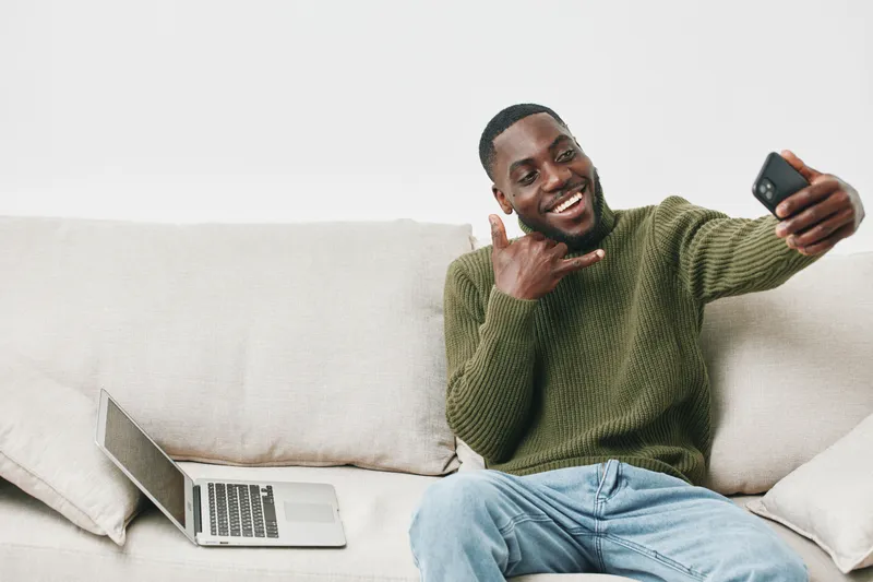 Happy young Black man taking a selfie on a couch, wearing a cozy green sweater, with a laptop nearby, reflecting a relaxed home atmosphere