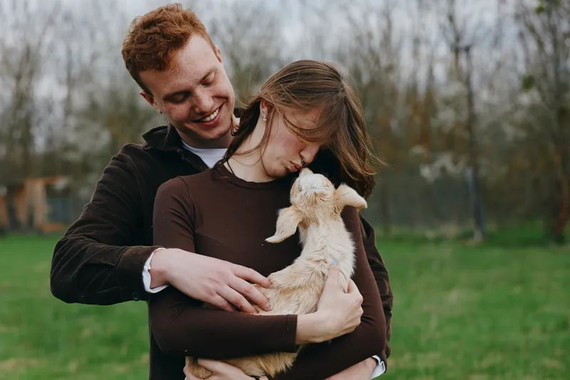 Couple hugging outdoors with a goat kid in a countryside setting, highlighting affection, love, and a wholesome lifestyle moment of bonding and care, with bright natural lighting.