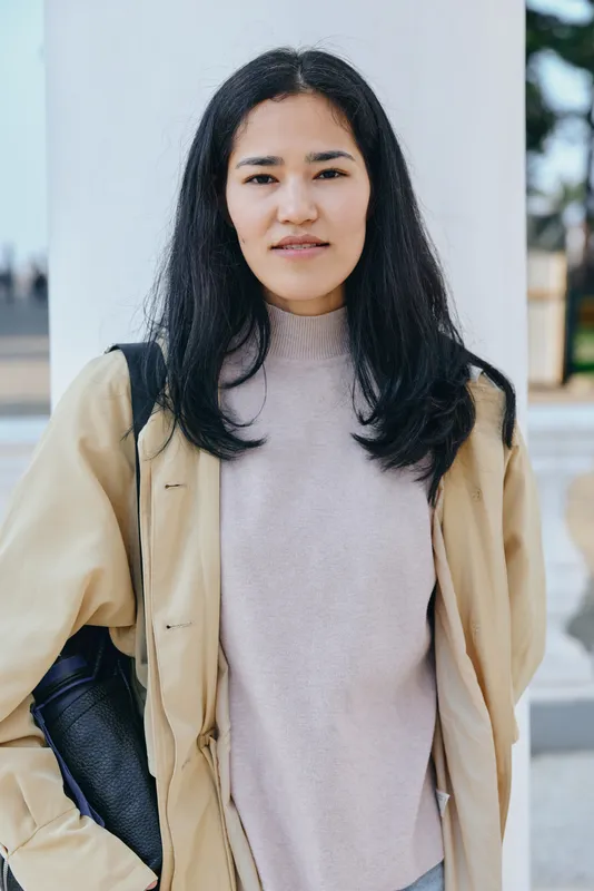 Woman in beige coat and soft pink sweater stands outdoors with a calm expression, urban backdrop, natural light, fashion photography, casual street style, everyday outfit, confident presence